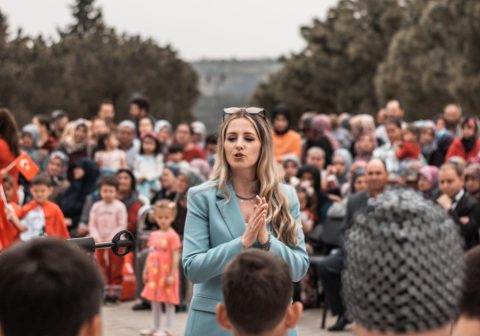 A woman addresses a large crowd during an outdoor community event, engaging with the audience.